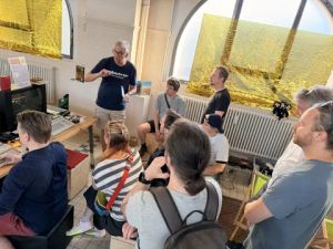 Vortragender vor Zuhörern in Raum mit goldener Fensterfolie und Vintage-Computertechnik.