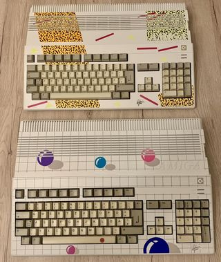 Draufsicht auf zwei originale Amiga 500 der „New Art Edition", die auf einem hellen Holzboden liegen. Das obere Gerät zeigt das „Leopard-Design" mit gelb-schwarzen Mustern und pinken Streifen. Das untere Gerät zeigt das „Ball-Design" mit bunten Kugeln auf einem grauen Gitter-Hintergrund. Beide Gehäuse tragen unten rechts die aufgedruckte Unterschrift von Stefanie Tücking.
